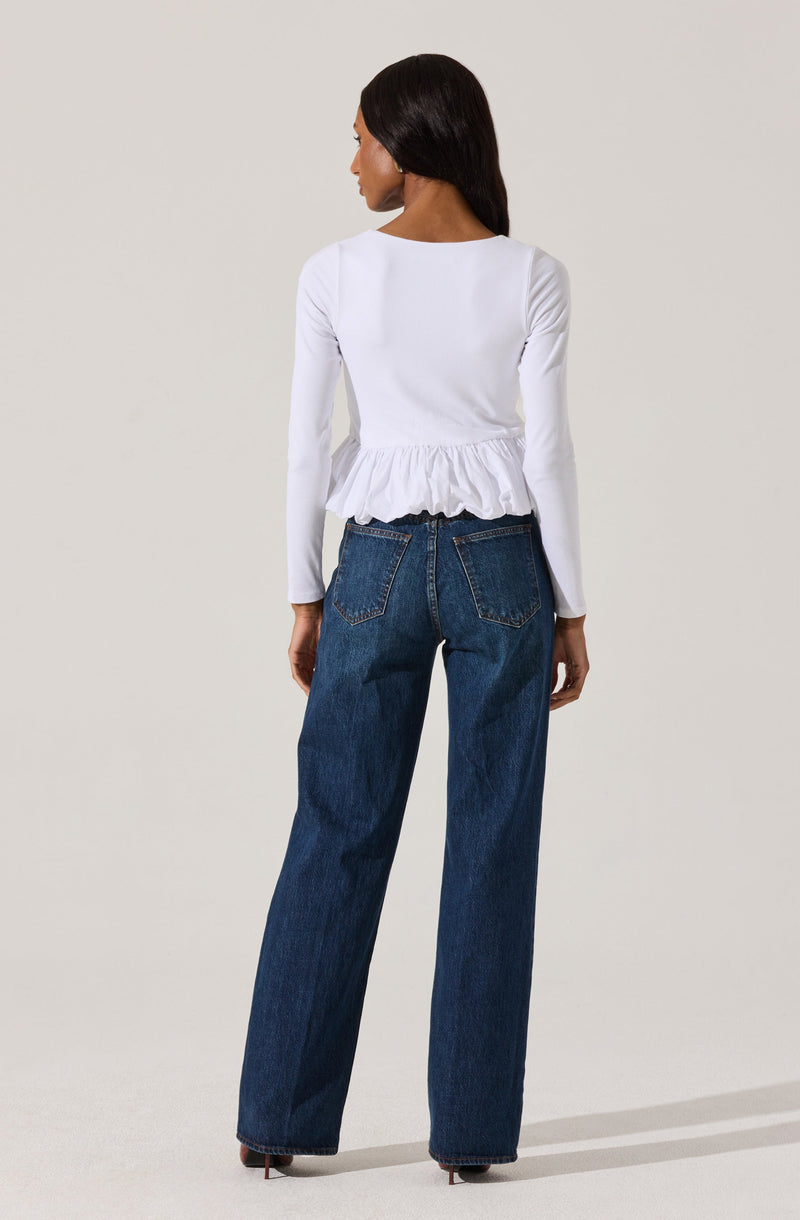 A woman is wearing a white long-sleeve top with a ruffled hem and a pair of high-waisted, wide-leg blue jeans. The top has a simple round neckline and the jeans have visible back pockets with stitching details.