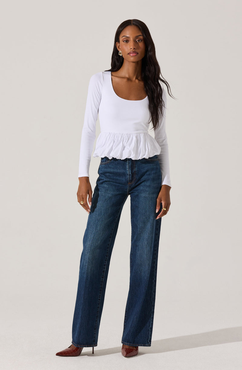A woman is wearing a white long-sleeve top with a scoop neckline and a ruffled hem, paired with high-waisted, straight-leg blue jeans. She completes the look with pointed-toe heels and gold earrings.