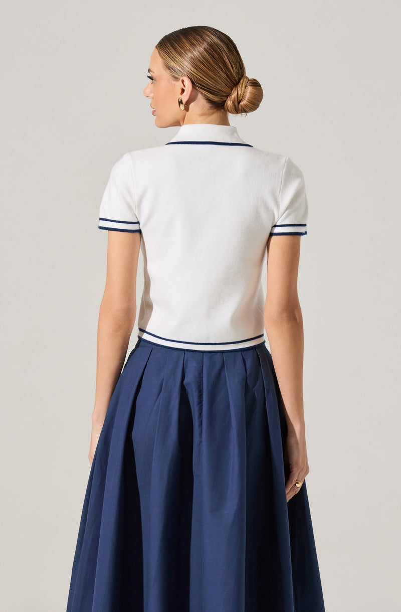A woman is wearing a white short-sleeved top with dark blue trim on the collar and sleeve cuffs, paired with a high-waisted, dark blue pleated skirt.