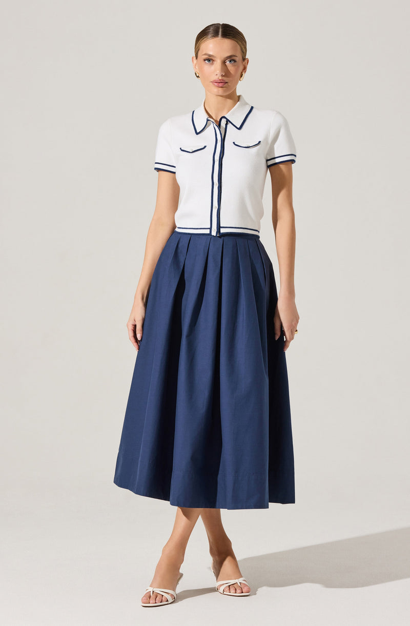 A woman is wearing a white short-sleeved knit top with navy piping details along the collar, sleeves, and front buttons. She pairs it with a high-waisted, full-length navy pleated skirt. She is also wearing white open-toe heels.