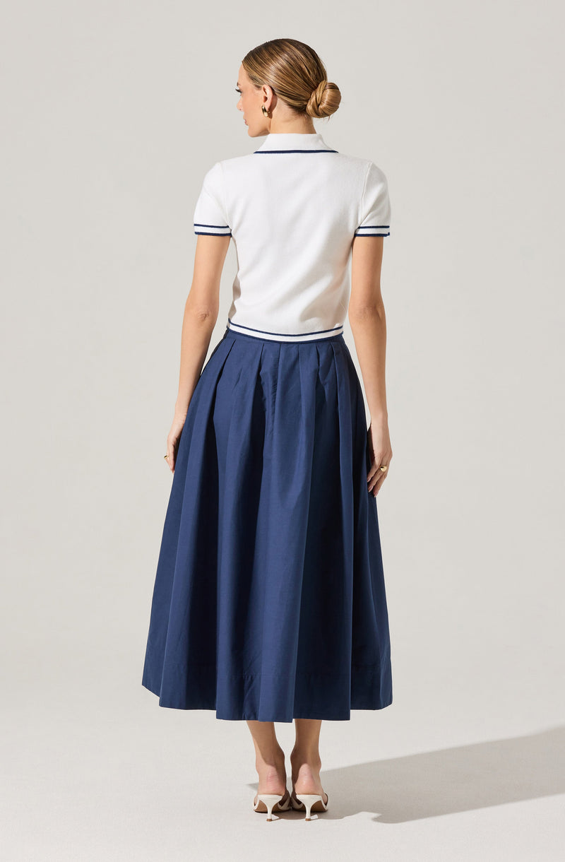 A woman is wearing a white short-sleeved polo shirt with dark blue trim on the collar and sleeves, paired with a high-waisted, dark blue, pleated, ankle-length skirt. She is also wearing nude-colored high-heeled shoes.