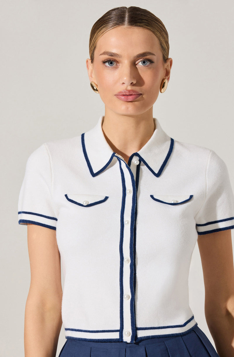 A woman is wearing a short-sleeved, white knit top with navy blue trim outlining the collar, front buttons, pockets, and sleeve cuffs. The top features two chest pockets with curved navy blue piping. She is also wearing gold hoop earrings.