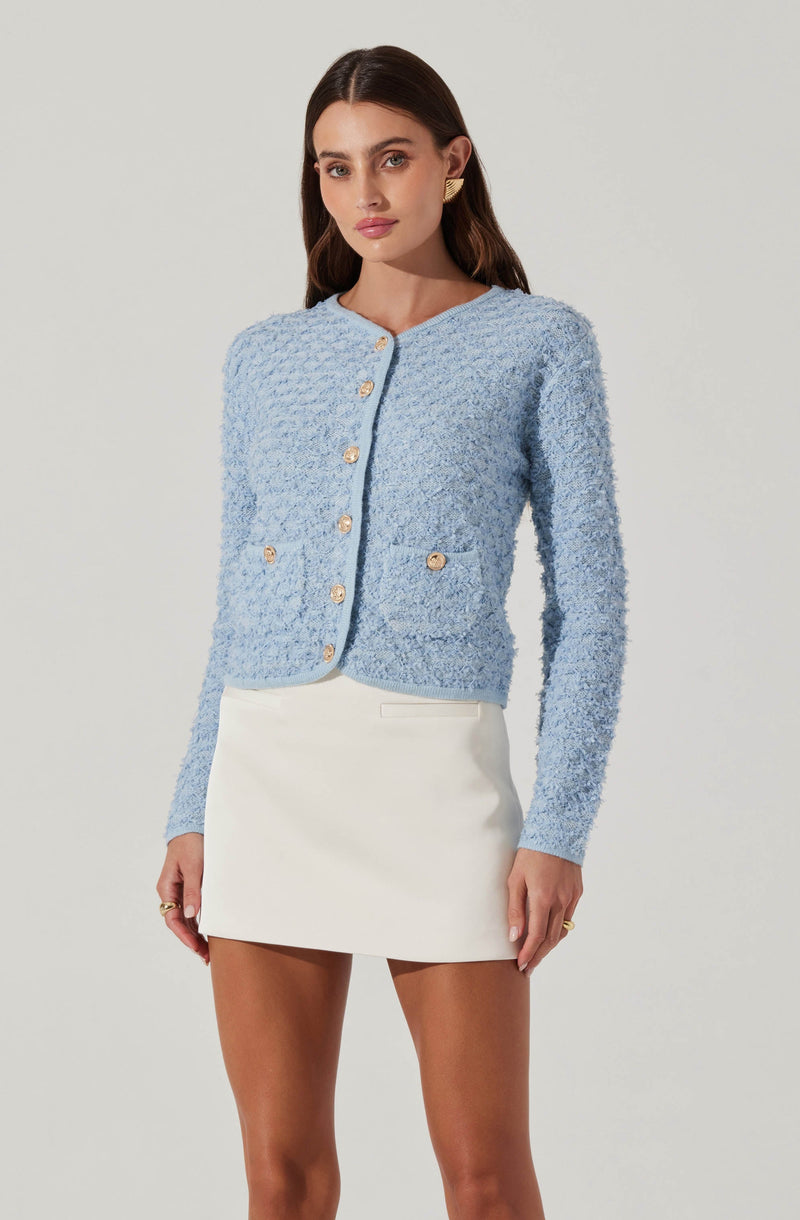 A woman is wearing a light blue, textured, long-sleeve cardigan with gold buttons down the front and on two front pockets. She pairs it with a white, high-waisted, A-line mini skirt.