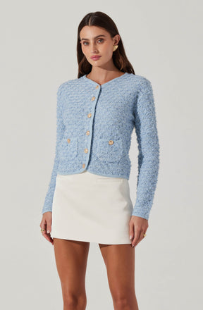 A woman is wearing a light blue, textured, long-sleeve cardigan with gold buttons down the front and on two front pockets. She pairs it with a white, high-waisted, A-line mini skirt.