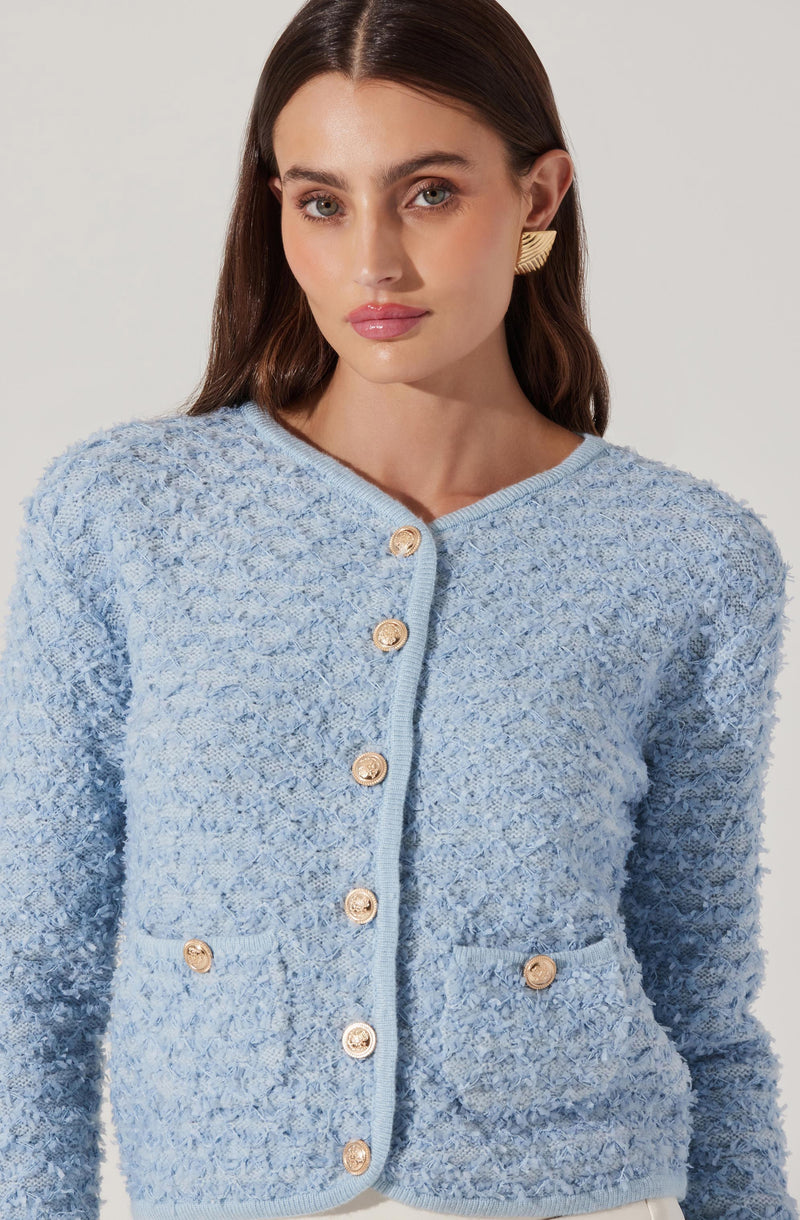 A woman is wearing a light blue, textured, boucle-style cardigan with gold buttons down the front. The cardigan features a rounded neckline and has two small pockets near the waist, each with a gold button. She is also wearing gold statement earrings.