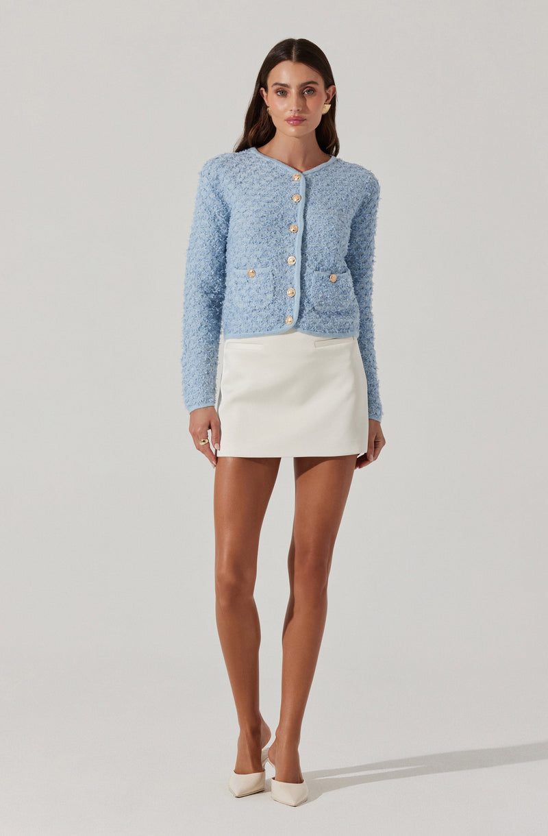 A woman is wearing a light blue, textured, long-sleeve cardigan with a round neckline, decorated with gold buttons down the front and on the pockets. She pairs the cardigan with a short, white, high-waisted skirt and white high-heeled shoes.