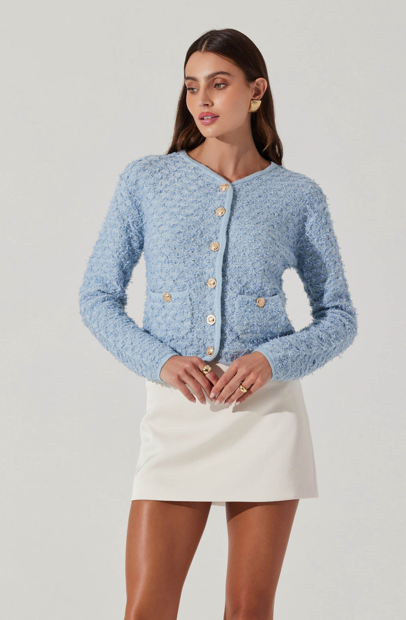 A woman is wearing a light blue, textured, button-up cardigan with gold buttons and two front pockets. She pairs it with a white mini skirt.