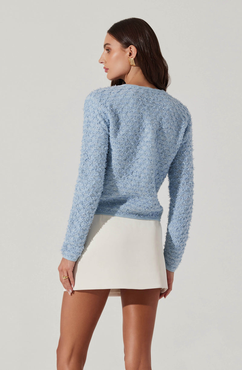 A woman is wearing a light blue textured long-sleeve sweater with a round neckline, paired with a high-waisted white mini skirt. She has dark brown hair and is accessorized with gold earrings and a gold ring.