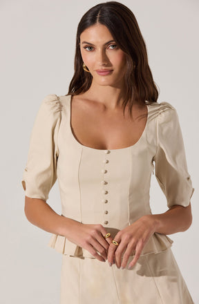 A woman is wearing a cream-colored top with puffed sleeves that have small cutouts and button detailing down the front. The top has a square neckline and a fitted waist with a peplum hem. She is accessorizing with gold hoop earrings and multiple gold rings.