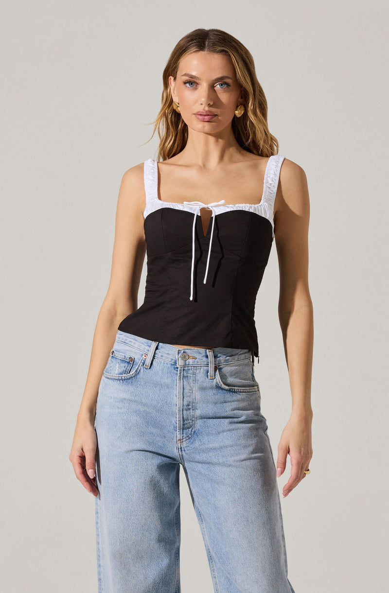 A woman is wearing a sleeveless top with wide white straps and a black bodice featuring a front keyhole cutout tied with a white string. She pairs the top with high-waisted, light-wash jeans.