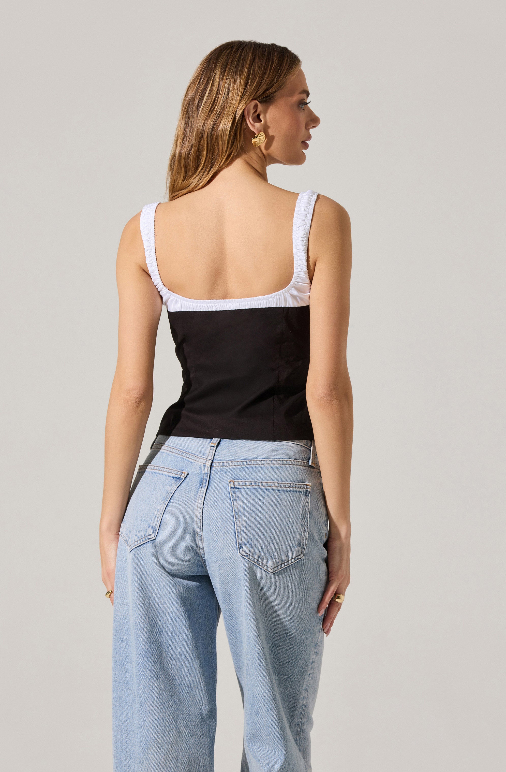 A woman is wearing a sleeveless top with wide white straps and a black body. The top has a square neckline and a fitted silhouette. She is also wearing light blue, high-waisted jeans with a relaxed fit.