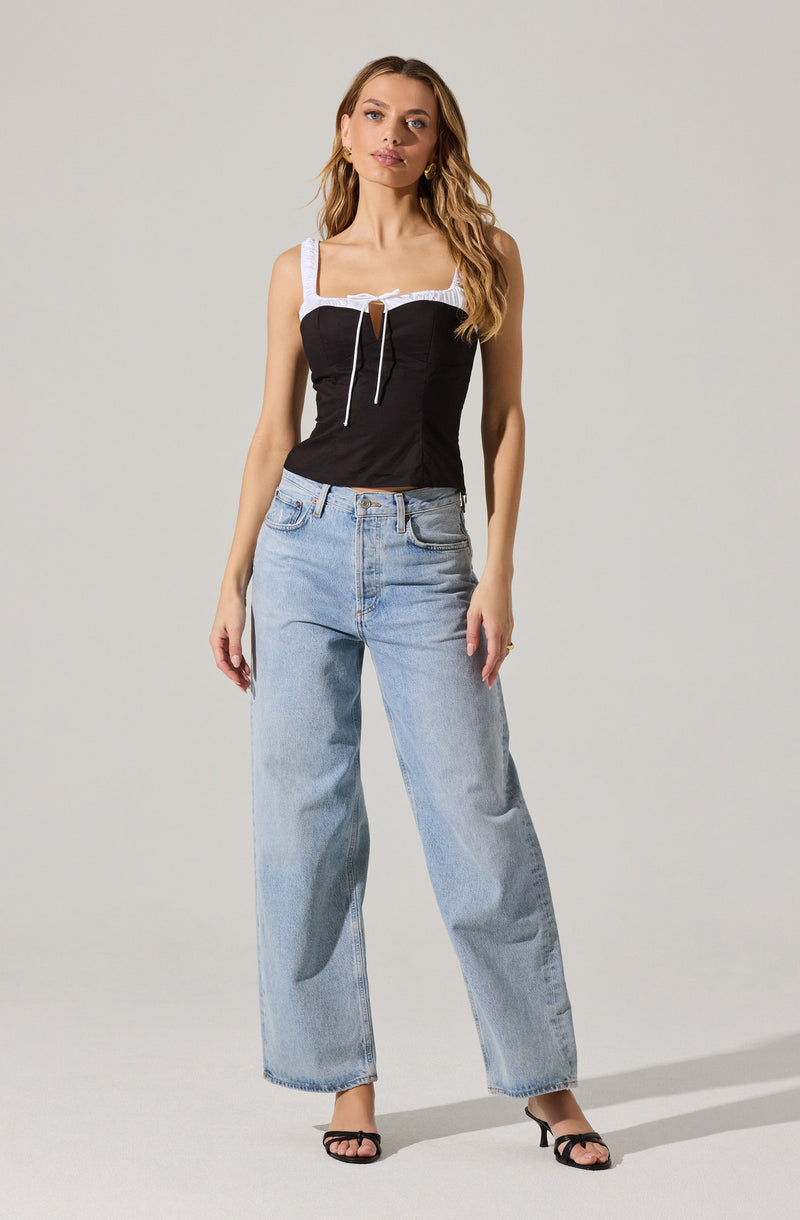 A woman is wearing a sleeveless, fitted black top with white lace accents on the straps and across the chest, featuring a small keyhole cutout at the center with a white drawstring tie. She is also dressed in high-waisted, light-wash, wide-leg jeans and black open-toe high-heeled sandals.