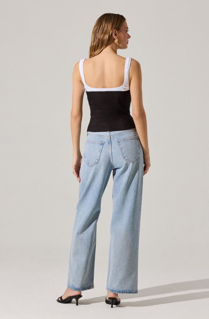 A woman is wearing a sleeveless top with wide straps and a square neckline. The top is black with white straps and trim along the neckline and armholes. She is also wearing light blue, high-waisted, wide-leg jeans with visible stitching and back pockets, and black heeled shoes.