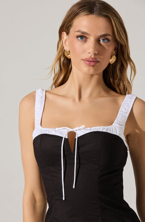 A woman is wearing a sleeveless dress with a fitted black bodice and white straps. The dress features a keyhole cutout at the chest, tied with a small bow, and has white trim along the straps and neckline. She is accessorized with gold, shell-shaped earrings.