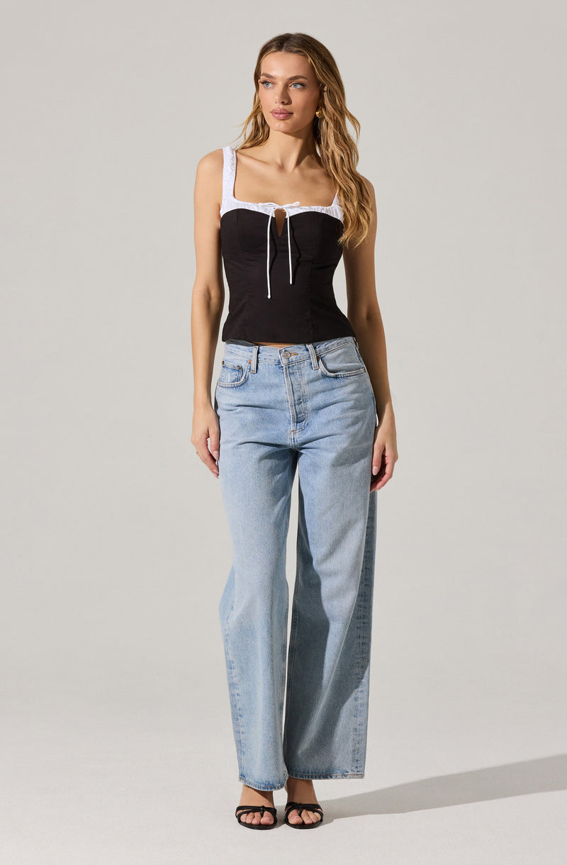 A woman is wearing a fitted black sleeveless top with white lace straps and a white lace trim at the neckline, featuring a keyhole cutout with a white tie detail in the front. She is also wearing high-waisted, light-wash, wide-leg jeans and black strappy high-heeled sandals.