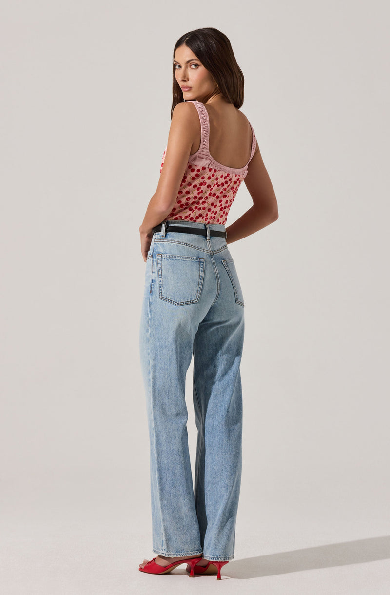 A woman is wearing a sleeveless pink top with red polka dots and lace trim around the armholes, paired with high-waisted, light-wash denim jeans that have large back pockets. She is also wearing red high-heeled shoes with a strap.