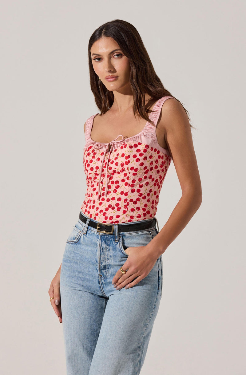A woman is wearing a sleeveless pink top with a cherry print design and a small bow at the neckline. She pairs it with light blue high-waisted jeans and a black belt.