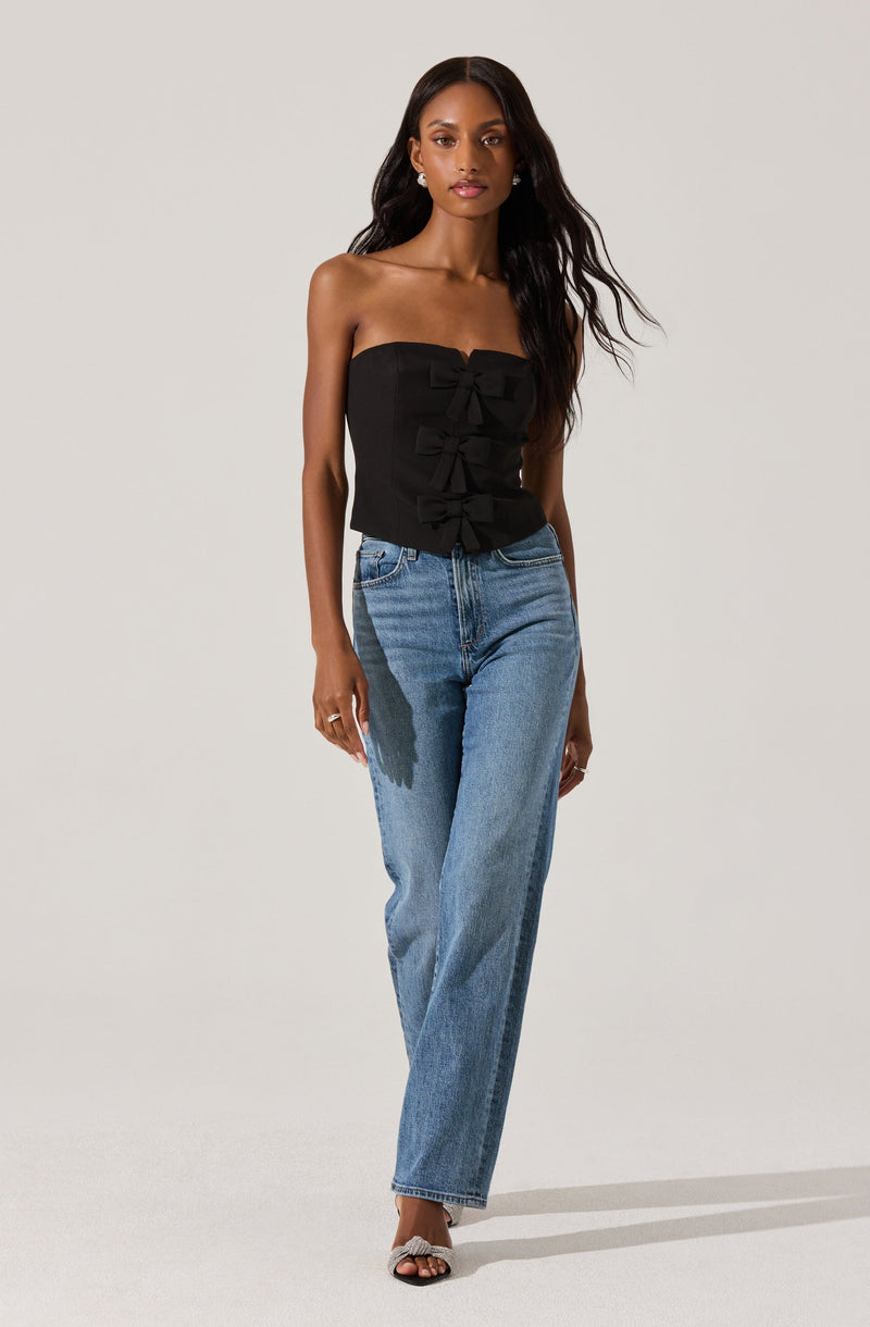 A woman is wearing a black strapless top with three decorative bows down the front and high-waisted, light-wash, straight-leg jeans. She is also wearing open-toe black and white sandals with a small heel.
