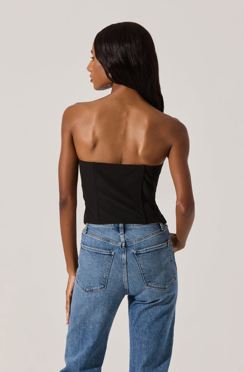 A woman is wearing a strapless black top and high-waisted blue jeans. The top is fitted and sleeveless, exposing her shoulders and back. The jeans are classic blue denim with visible stitching and back pockets. She has long, straight, dark hair.