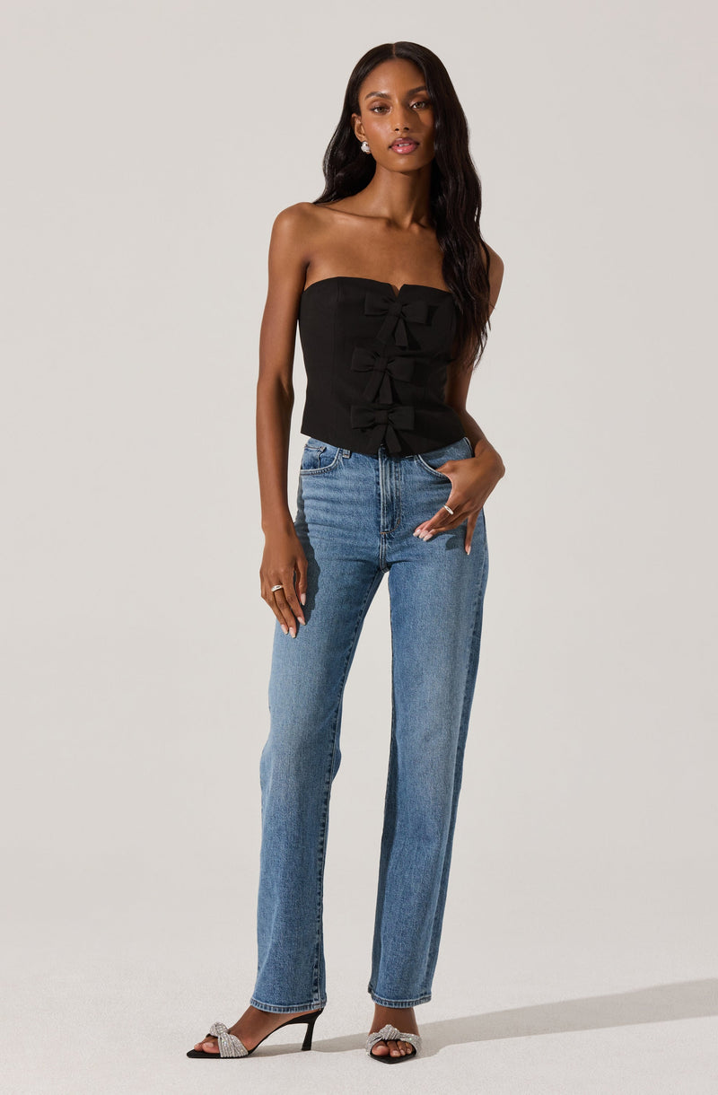 A woman is wearing a strapless black top with ruffled details down the front, paired with high-waisted blue jeans. She is also wearing open-toe high heels with a decorative bow on the front.