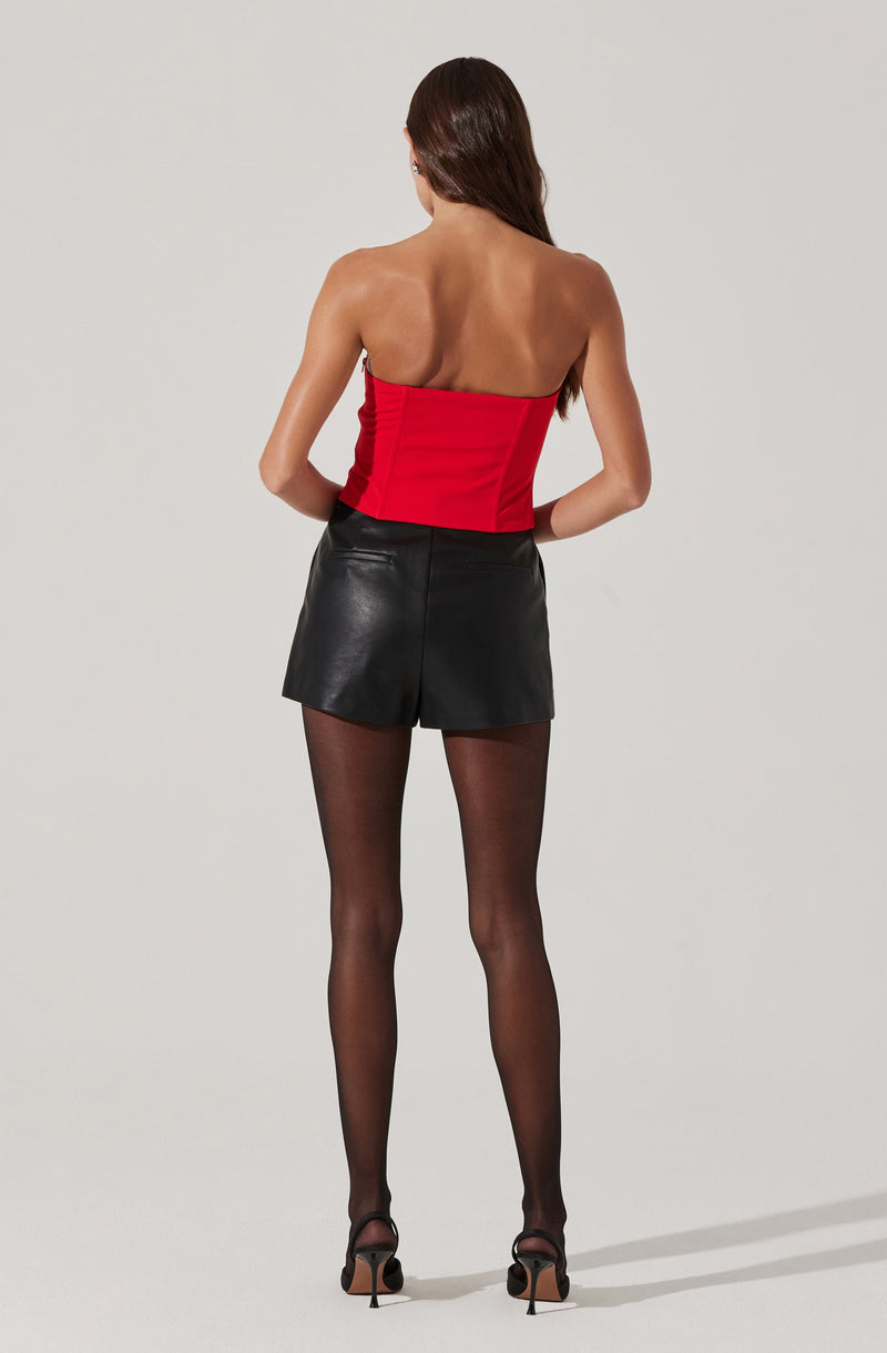 A woman is wearing a strapless, bright red corset-style top and high-waisted black leather shorts. She is also wearing sheer black stockings and black high-heeled shoes. Her long, dark hair is worn down, and she is facing away from the camera.