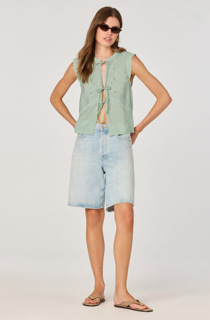 A woman is wearing a sleeveless, light green embroidered top with a front opening tied with a bow, paired with light-wash denim shorts that reach just above the knees. She accessorizes with dark sunglasses and gold hoop earrings, and she is wearing flat sandals.