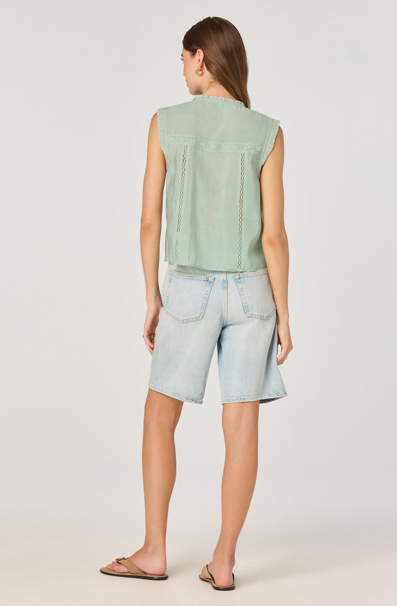 A woman is wearing a sleeveless, light green top with decorative stitching and lace details. She pairs it with light-wash denim shorts that are knee-length and have large back pockets. She is also wearing tan flat sandals.
