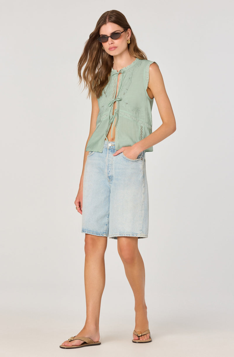 A woman is wearing a sleeveless, light green, button-up top with a tie detail at the front and embroidered accents. She pairs it with light-washed, knee-length denim shorts. She accessorizes with dark sunglasses and gold earrings, and she is wearing tan sandals.