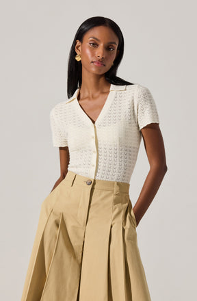 A woman is wearing a short-sleeved, cream-colored knitted top with a subtle V-neckline and buttons down the front. She pairs it with high-waisted, beige wide-leg pants that feature pleats and large front pockets. She accessorizes with gold hoop earrings.