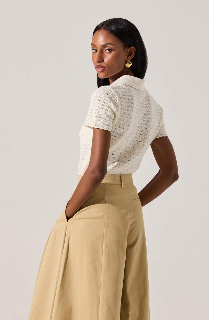 A woman is wearing a short-sleeved, cream-colored, textured knit top with a collar. She has paired it with high-waisted, wide-leg beige trousers. The outfit is accessorized with large, gold, leaf-shaped earrings.