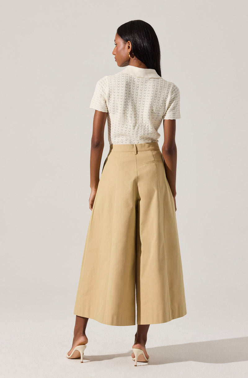 A woman is wearing a short-sleeved, textured white knit top with a collar, tucked into high-waisted, wide-leg beige pants that reach just above her ankles. She is also wearing white high-heeled shoes.