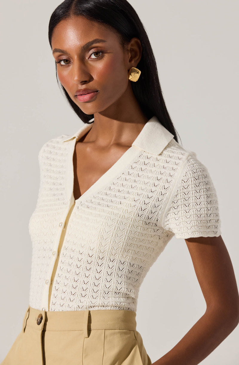 A woman wearing a short-sleeved, knit, cream-colored blouse with a collar and button-down front. The fabric has a delicate, textured, open-knit pattern. She is paired with high-waisted beige pants.