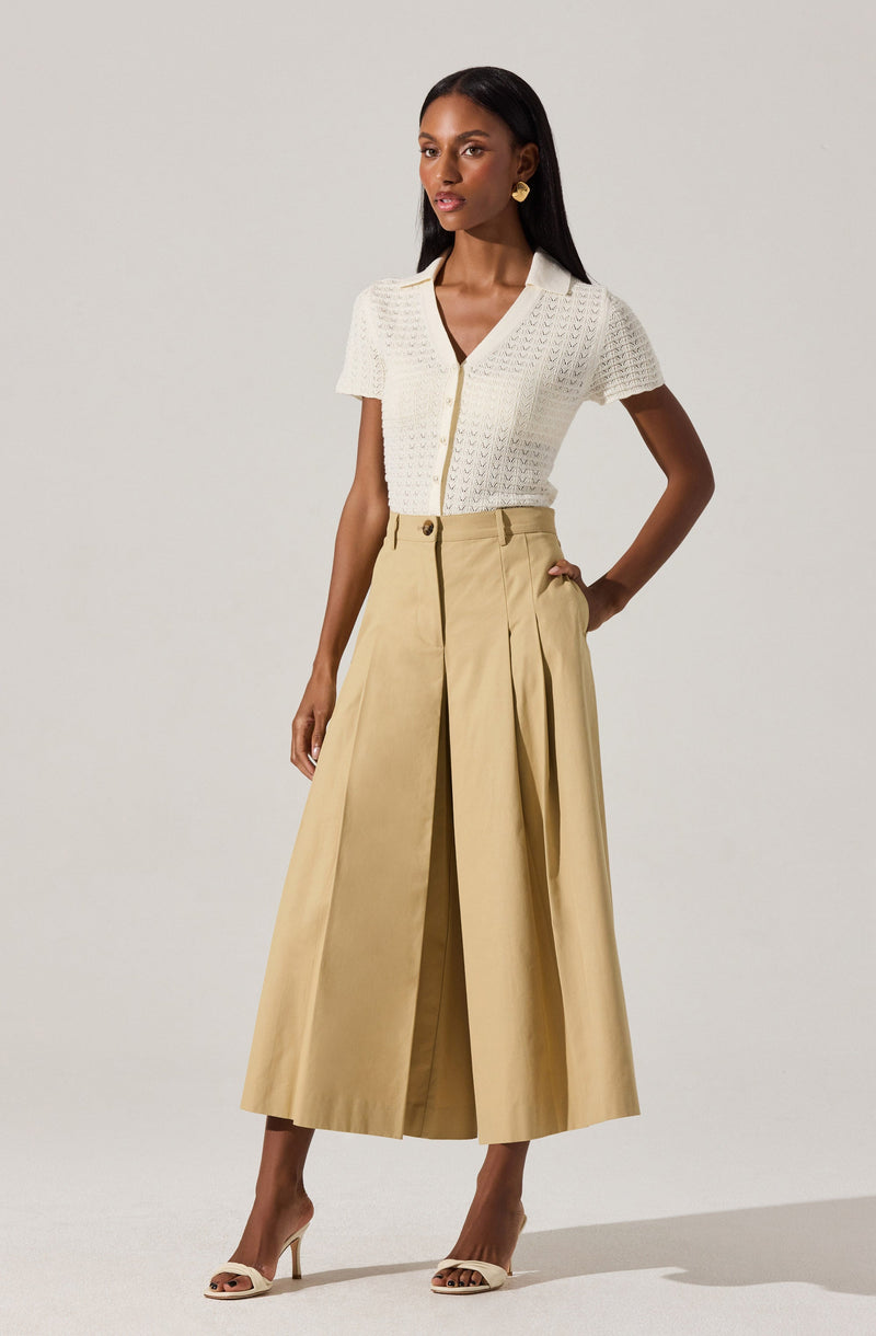 A woman is wearing a white short-sleeved, textured knit top with a collared neckline and a button-down front. She has paired the top with high-waisted, wide-leg beige culottes that feature pleats and a front zipper with a button closure. She is also wearing beige high-heeled sandals and gold earrings.