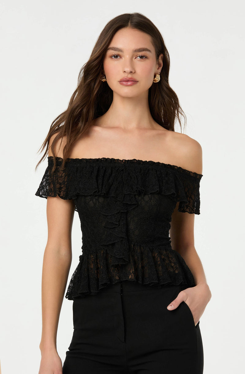 A woman is wearing an off-the-shoulder black lace top with ruffled details around the neckline, sleeves, and hem. The top has a fitted silhouette and features ruffles down the front. She pairs it with black pants and accessorizes with gold hoop earrings.