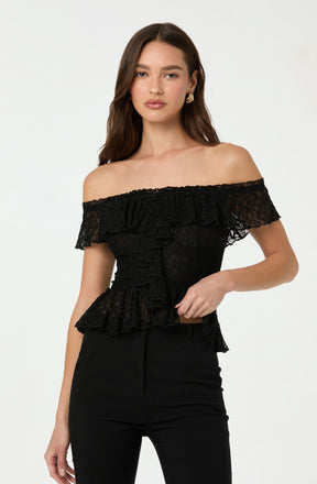A woman is wearing an off-the-shoulder black lace top with ruffle details. The top features a floral lace pattern and has a layered ruffle design along the neckline and hem. She pairs it with high-waisted black pants.