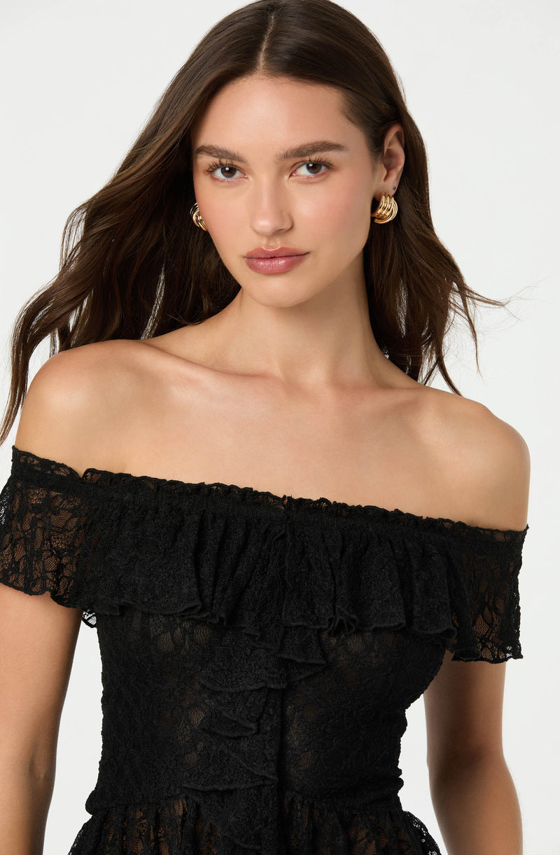 A woman with long, dark, wavy hair is wearing an off-the-shoulder black lace dress with a ruffled neckline. She has gold hoop earrings and a neutral expression on her face.