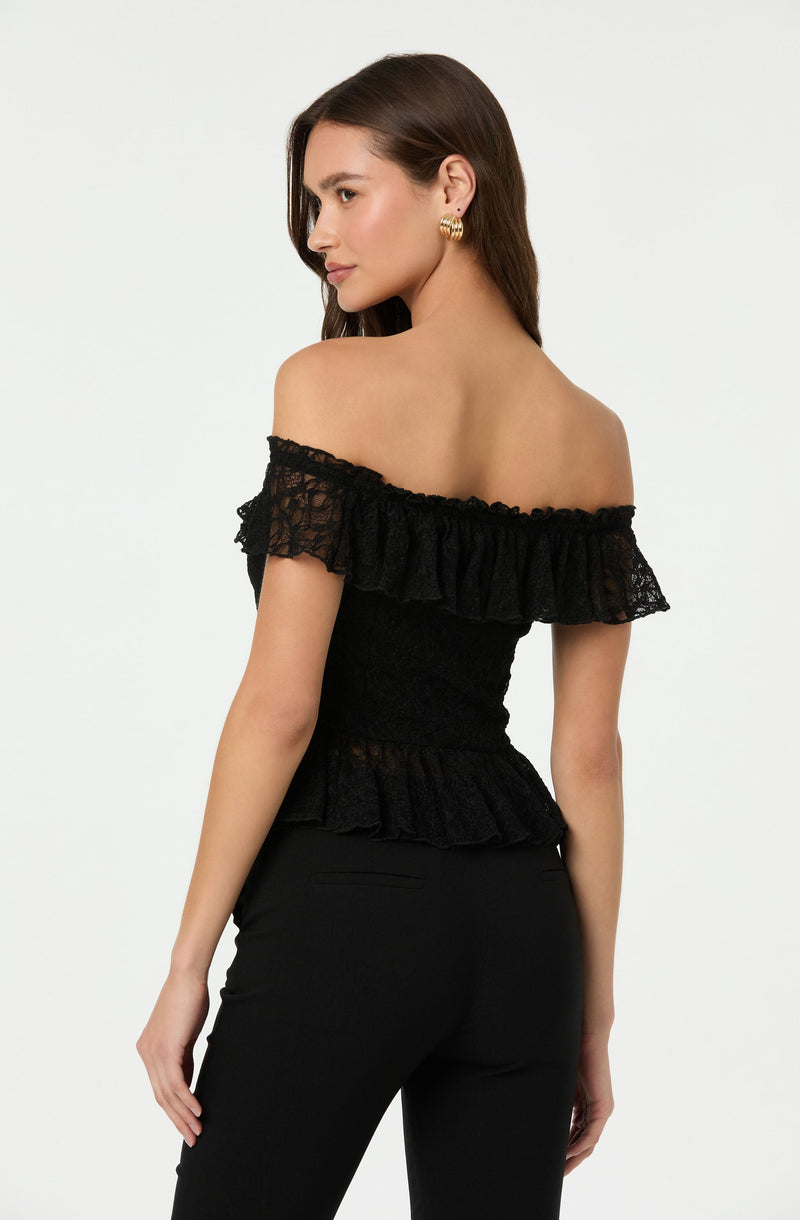 The woman is wearing an off-the-shoulder black top with ruffled lace detailing around the neckline and waist. The top has a fitted waist and the ruffles create a layered effect. She is also wearing black pants.