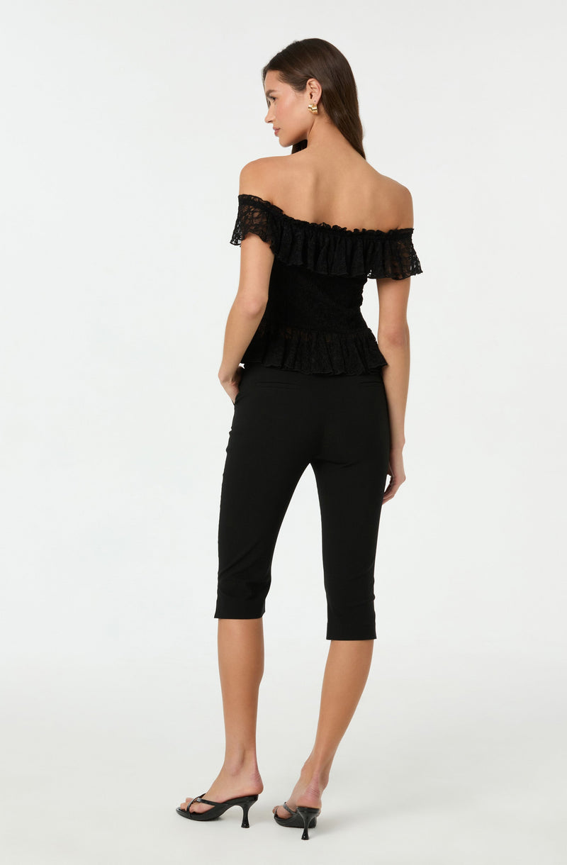 A woman is wearing an off-the-shoulder black top with ruffled lace detailing along the neckline and hem. She pairs it with fitted black cropped pants and black high-heeled sandals.