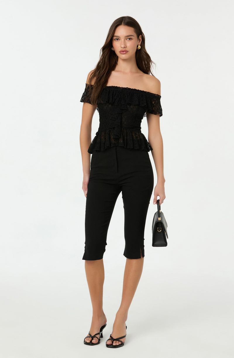 A woman is wearing an off-the-shoulder black lace top with ruffled detailing around the neckline and waist, paired with black cropped pants that have a small slit at the hem. She is also holding a small black handbag and wearing black strappy high-heeled sandals.