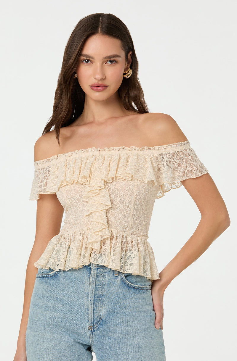 A woman is wearing an off-the-shoulder top made of beige lace fabric. The top features ruffles along the neckline, a ruffle at the waistline, and an additional ruffle detail down the front. The lace pattern is intricate and floral. She pairs the top with light-wash blue jeans.