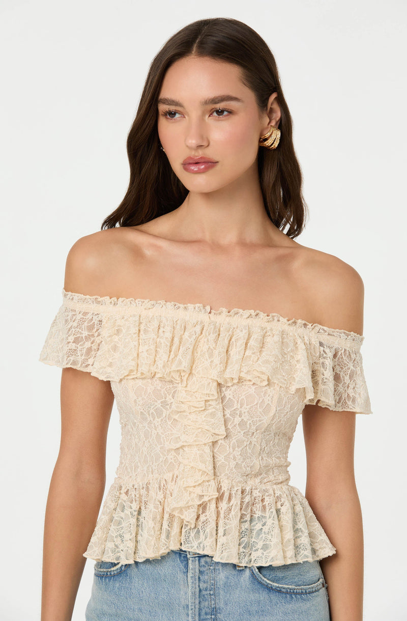 A woman is wearing an off-the-shoulder, cream-colored lace top with ruffled detailing along the neckline, shoulders, and hem. The top has an intricate lace pattern and a ruffled front design.