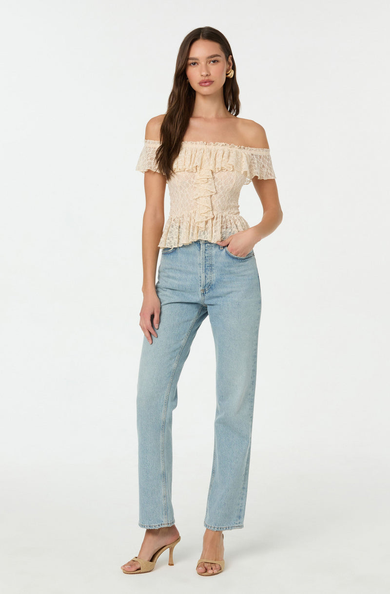 A woman is wearing an off-the-shoulder, cream-colored, ruffled top with a textured fabric. She has paired it with light blue high-waisted jeans and beige open-toe high heels.