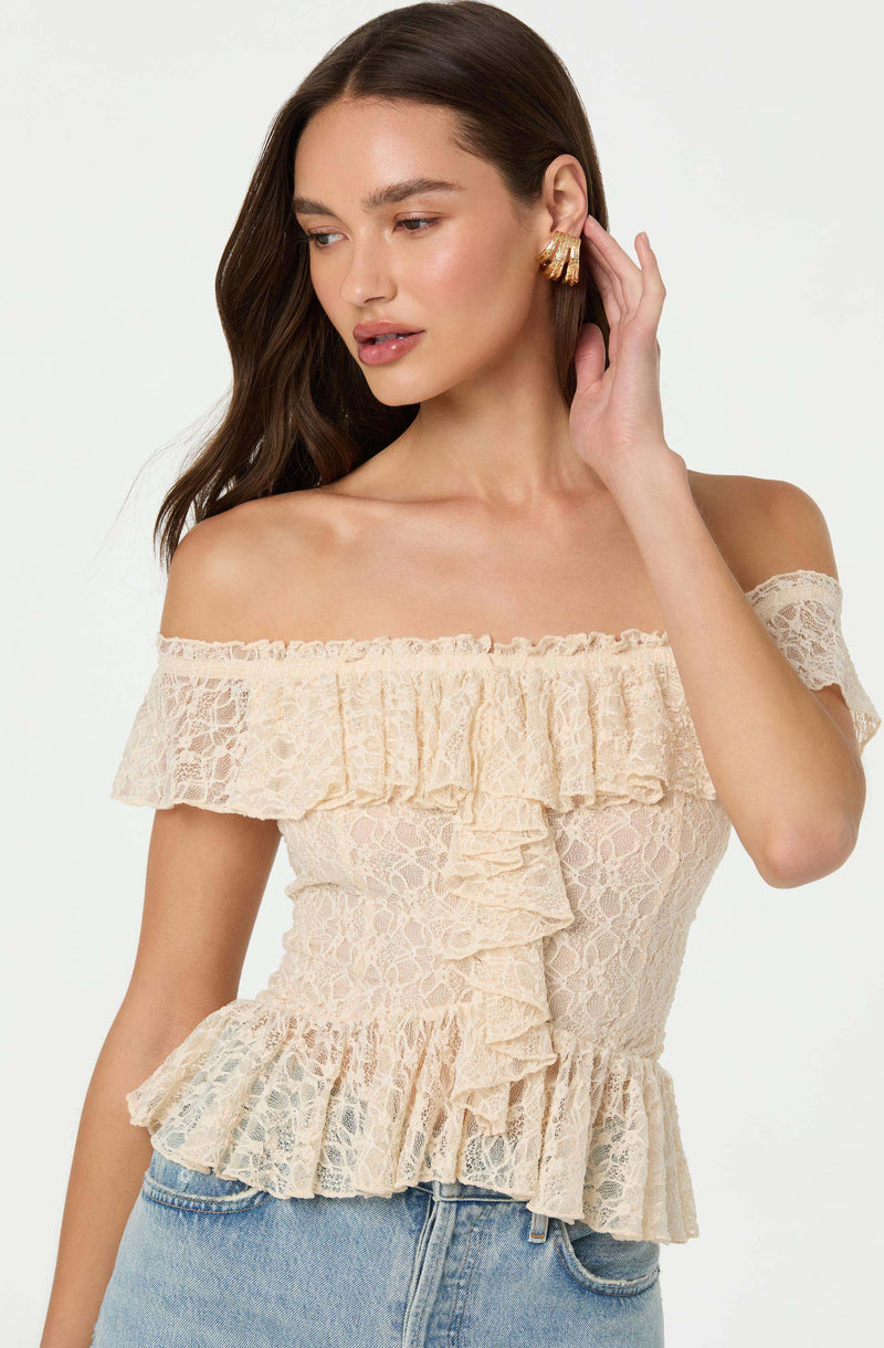 A woman is wearing an off-the-shoulder, cream-colored lace top with ruffles along the neckline, sleeves, and hem. The top has a fitted waist and a layered ruffle detail down the front.