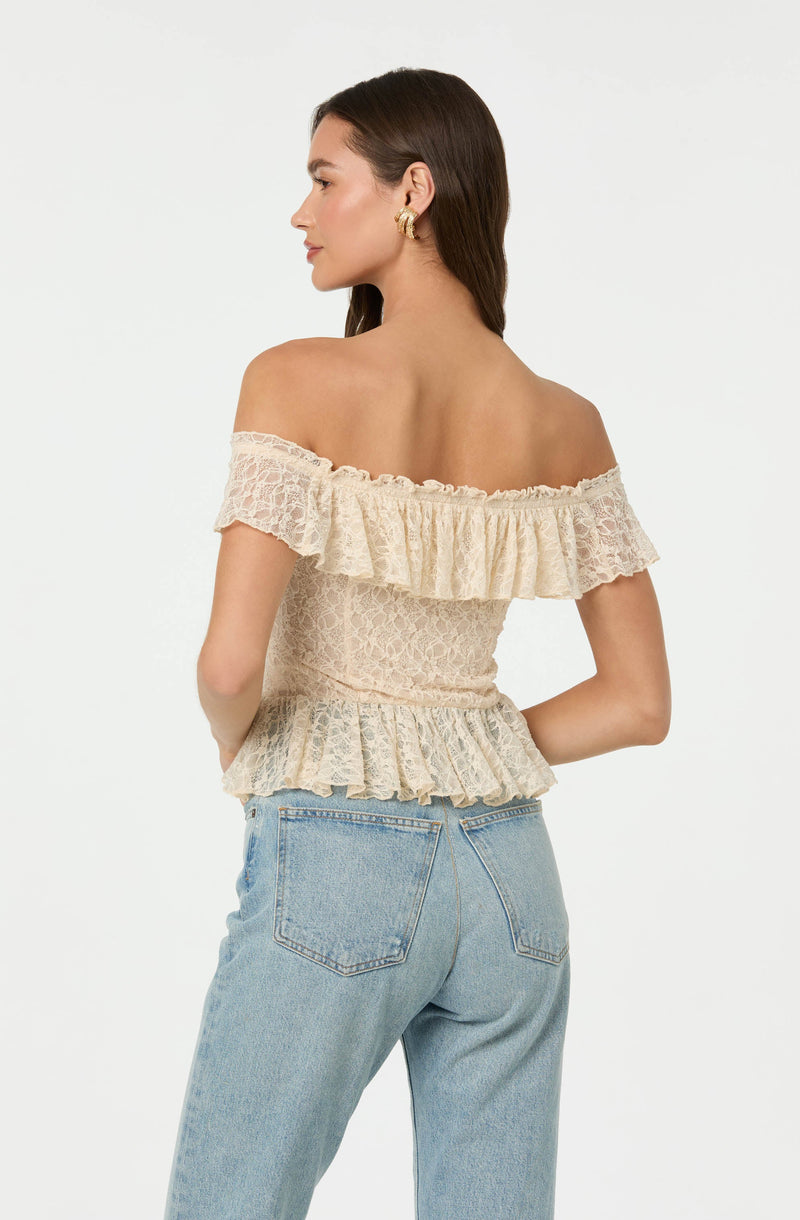 A woman is wearing an off-the-shoulder beige lace top with ruffled detailing along the neckline, waist, and sleeves. She pairs it with light blue high-waisted jeans.