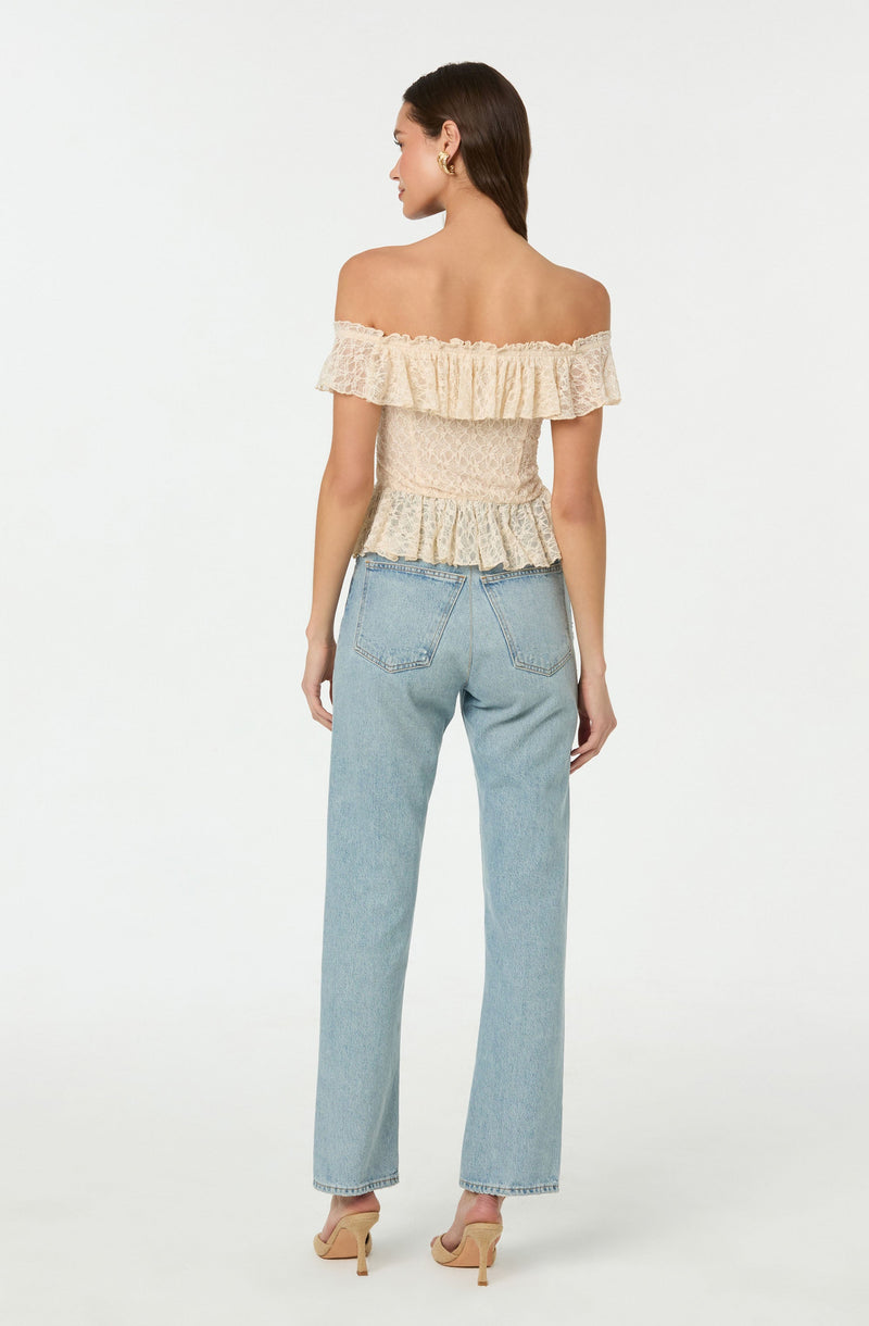 A woman is wearing an off-the-shoulder, cream-colored top with a ruffled neckline and textured, smocked fabric. She pairs it with light-wash, high-waisted, straight-leg jeans. She also wears beige high-heeled sandals and gold hoop earrings.