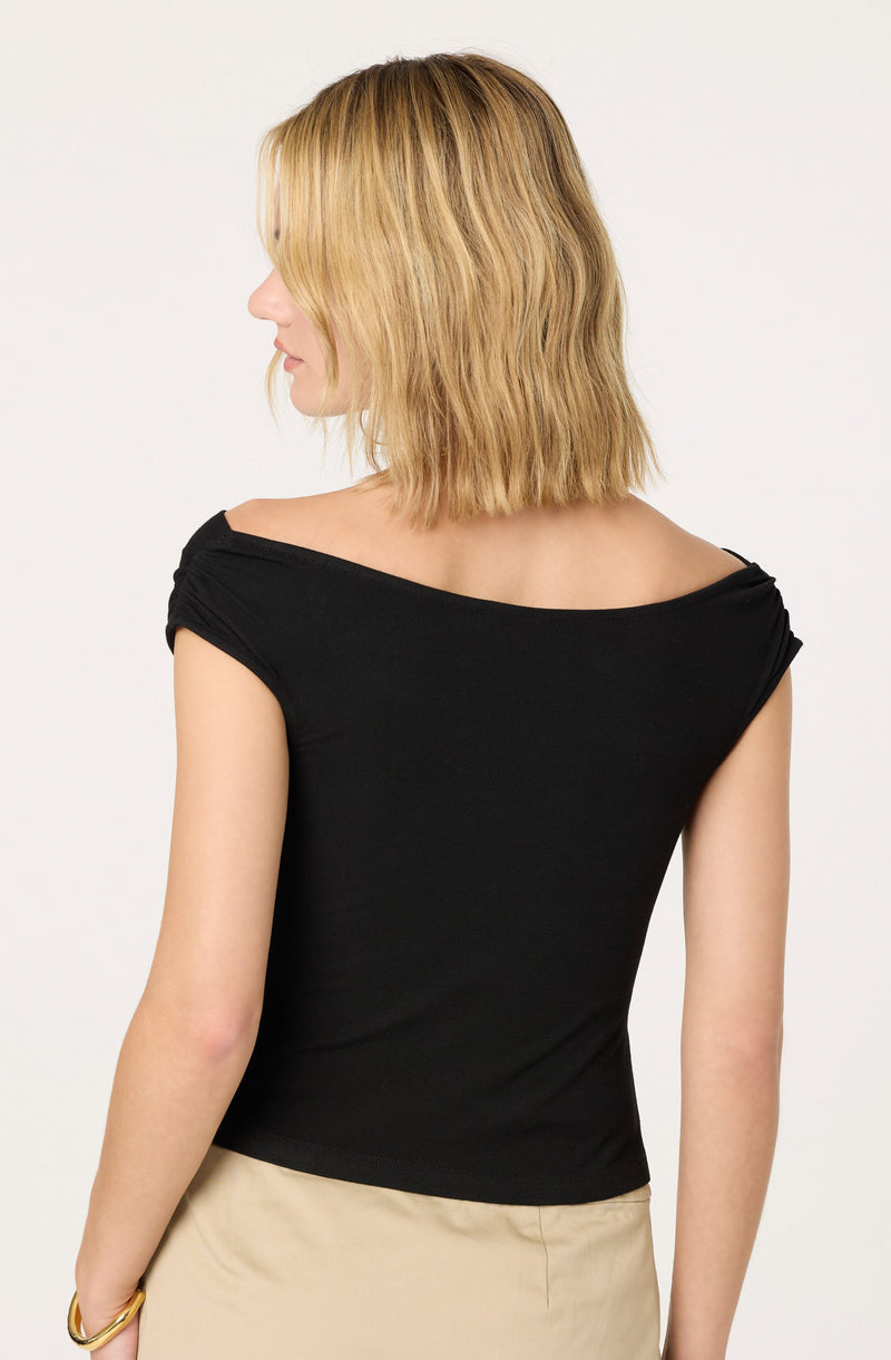 A woman is wearing a black, short-sleeved top with a wide, slightly off-the-shoulder neckline. The top fits snugly and is made of a smooth fabric. She is also wearing a beige skirt.