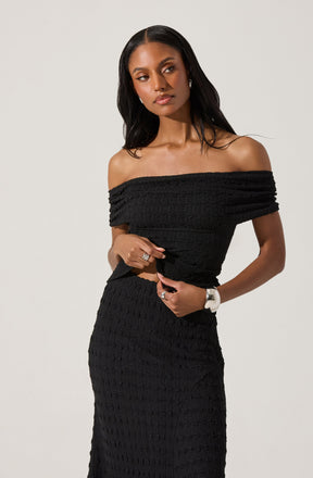 A woman is wearing an off-the-shoulder black dress made from textured fabric with a subtle grid pattern. The dress has a fitted silhouette, highlighting her waist and hips. She accessorizes with silver jewelry, including earrings and rings, and a white wristwatch.