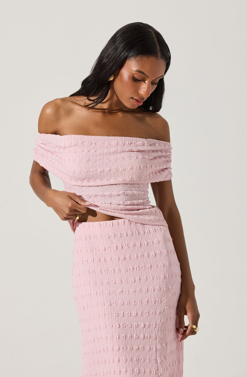 A woman is wearing a two-piece pink textured fabric outfit. The top is an off-the-shoulder style with a loose, relaxed fit, and the skirt is high-waisted and fitted, both made from the same fabric with a raised, dotted pattern.
