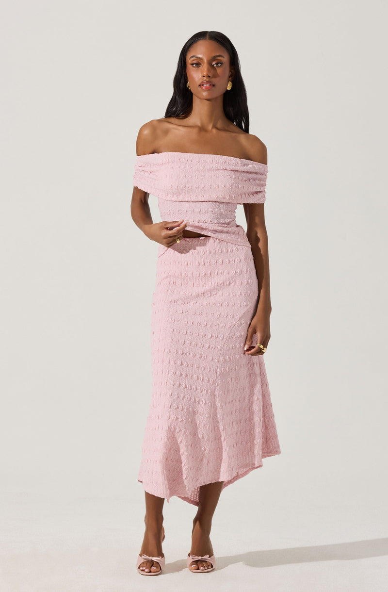 A woman is wearing a light pink, off-the-shoulder dress with a textured, crinkled fabric. The dress has a wide neckline that drapes around her shoulders and an asymmetrical hem that falls below her knees. She is also wearing matching light pink high-heeled sandals.