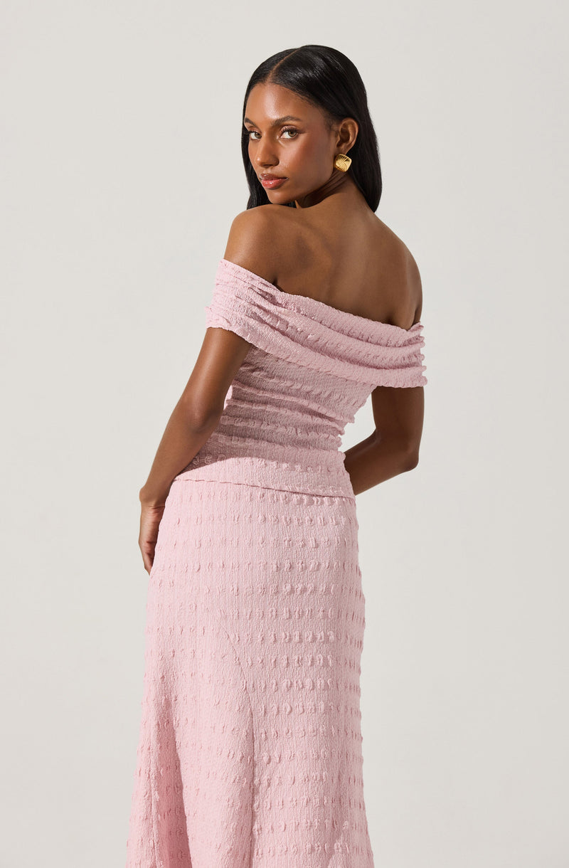 A woman is wearing a light pink dress with a textured, grid-like pattern. The dress has an off-the-shoulder neckline with wide, ruched fabric at the top that drapes over her shoulders and upper arms. The skirt of the dress is long and fitted, extending to the floor, with a consistent textured pattern throughout. She has long, straight black hair and is accessorized with large, round, gold earrings.